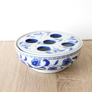 May include: A white and blue ceramic flower frog with a matching lid. The lid has six holes for arranging flowers. The bowl and lid are decorated with blue floral patterns. The bowl sits on a light wood surface.