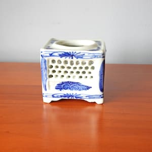 May include: A square, white ceramic incense burner with blue floral and geometric patterns. The burner has a circular opening at the top and a series of small holes on the sides. The design is reminiscent of traditional Asian pottery.