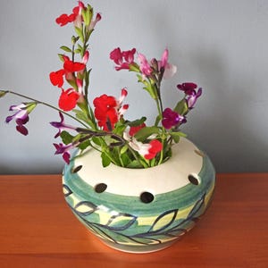 May include: A ceramic vase with a green and white floral design, featuring a ring of holes around the top. The vase is filled with a variety of colourful flowers, including red, pink, and purple.