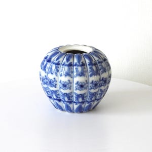 May include: A round, blue and white ceramic vase with a floral pattern. The vase has a scalloped rim and a dark interior. It is sitting on a white surface.