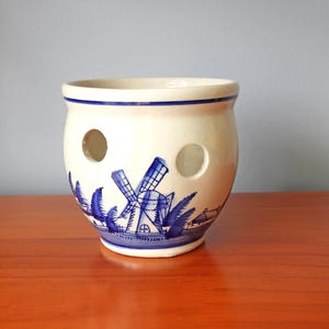 May include: Cream ceramic pot with a blue windmill and foliage design. The pot has two circular holes and a blue rim. The base of the pot has a blue border with a windmill and boat scene.