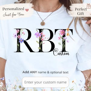 Puede incluir: Camiseta blanca con las letras "RBT" en negro, adornada con detalles florales en rosa, morado y amarillo. La palabra "Custom" está debajo de las letras. La camiseta también tiene el texto "Add ANY name & optional text" y "Enter your custom name".