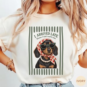 May include: An ivory-colored crewneck sweatshirt featuring a graphic of a black and tan dachshund wearing a red and white polka dot headscarf, green sunglasses, and a pearl necklace. The text reads I ARRIVED LATE Because I didn't want to come.