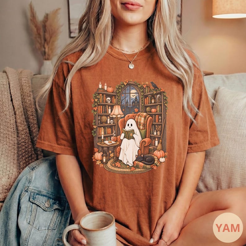 May include: A rust-colored short-sleeve t-shirt featuring a graphic print of a white ghost sitting in an armchair reading a book. The scene includes bookshelves, a window with a night sky, pumpkins, candles, and a black cat on a rug.