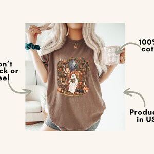 May include: A brown t-shirt featuring a graphic of a ghost sitting in an armchair reading a book in a library surrounded by bookshelves, pumpkins, and candles. Text on the image includes 100% US cotton, Won't Crack or Peel, and Produced in USA.
