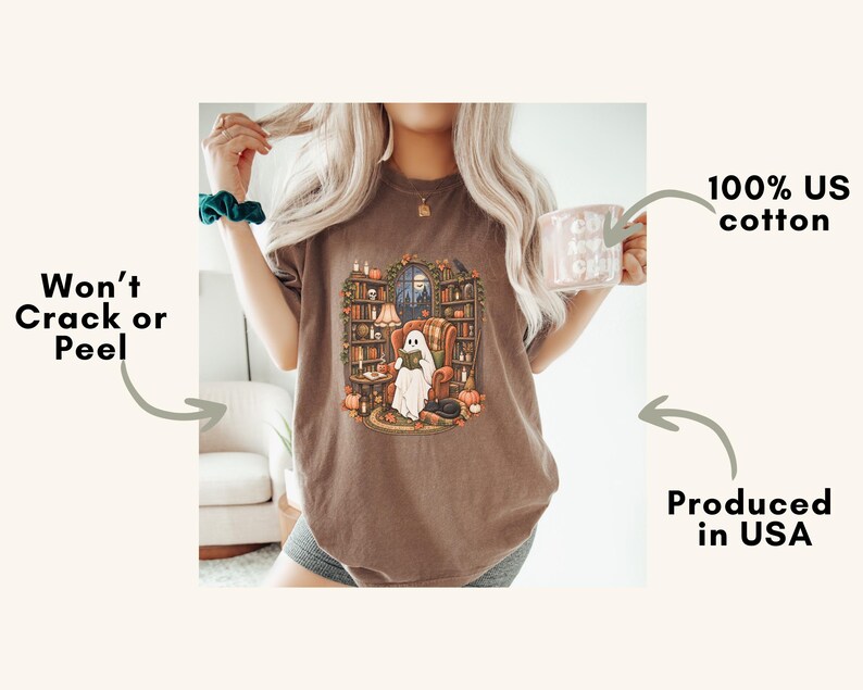 May include: A brown t-shirt featuring a graphic of a ghost sitting in an armchair reading a book in a library surrounded by bookshelves, pumpkins, and candles. Text on the image includes 100% US cotton, Won't Crack or Peel, and Produced in USA.