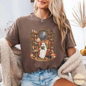 May include: A brown short-sleeve t-shirt featuring a graphic illustration of a white ghost sitting in an armchair reading a book. The scene includes bookshelves, pumpkins, candles, and a black cat, all set within a cozy library environment.