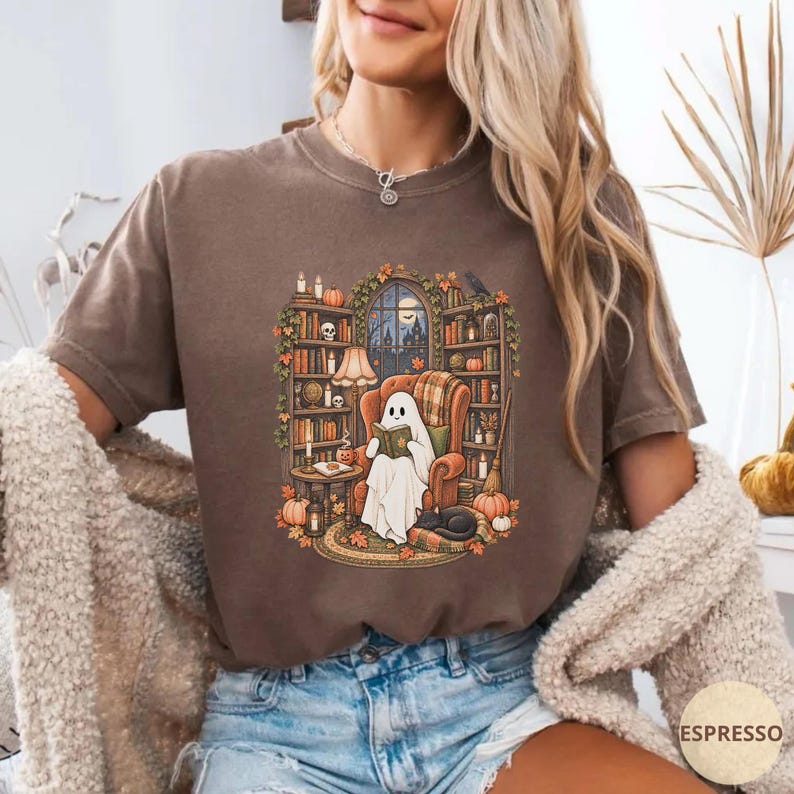 May include: A brown short-sleeve t-shirt featuring a graphic illustration of a white ghost sitting in an armchair reading a book. The scene includes bookshelves, pumpkins, candles, and a black cat, all set within a cozy library environment.