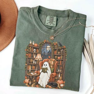 May include: A sage green crewneck t-shirt featuring a graphic print of a ghost reading a book in an armchair surrounded by bookshelves, pumpkins, and a black cat. The shirt has a Comfort Colors label at the neck and a thin gold chain necklace.