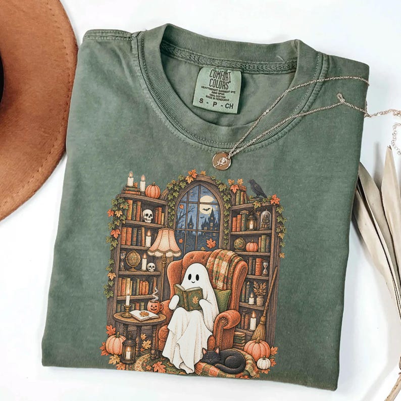 May include: A sage green crewneck t-shirt featuring a graphic print of a ghost reading a book in an armchair surrounded by bookshelves, pumpkins, and a black cat. The shirt has a Comfort Colors label at the neck and a thin gold chain necklace.