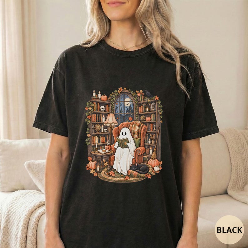 May include: A black short-sleeve t-shirt featuring a graphic print of a white ghost sitting in an orange armchair, reading a book. The scene includes bookshelves, pumpkins, candles, and a black cat, all framed by autumn leaves and a window view.