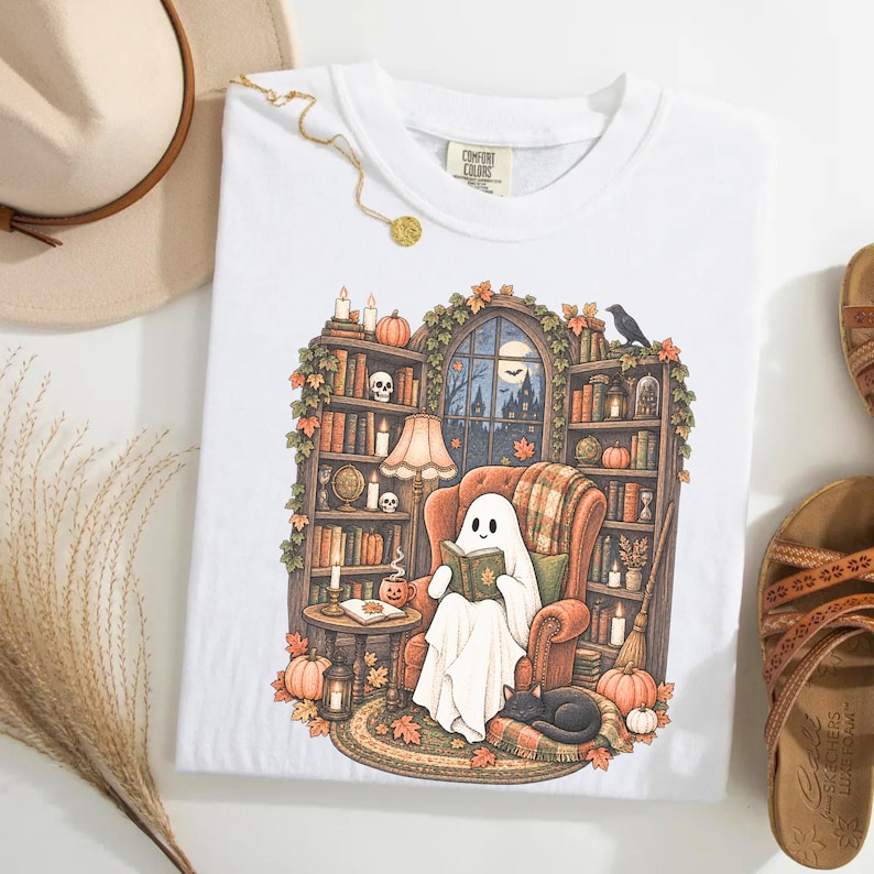 May include: A white t-shirt featuring a graphic illustration of a ghost sitting in an armchair reading a book. The scene includes bookshelves, a lamp, a black cat, pumpkins, and a window showing a night sky with a moon and a haunted house.