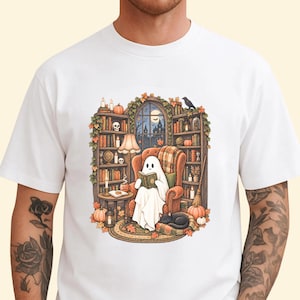 May include: A white t-shirt featuring a graphic illustration of a ghost sitting in an orange armchair, reading a book. The scene includes bookshelves, a lamp, a black cat, pumpkins, and autumn leaves, set against a window with a night view.