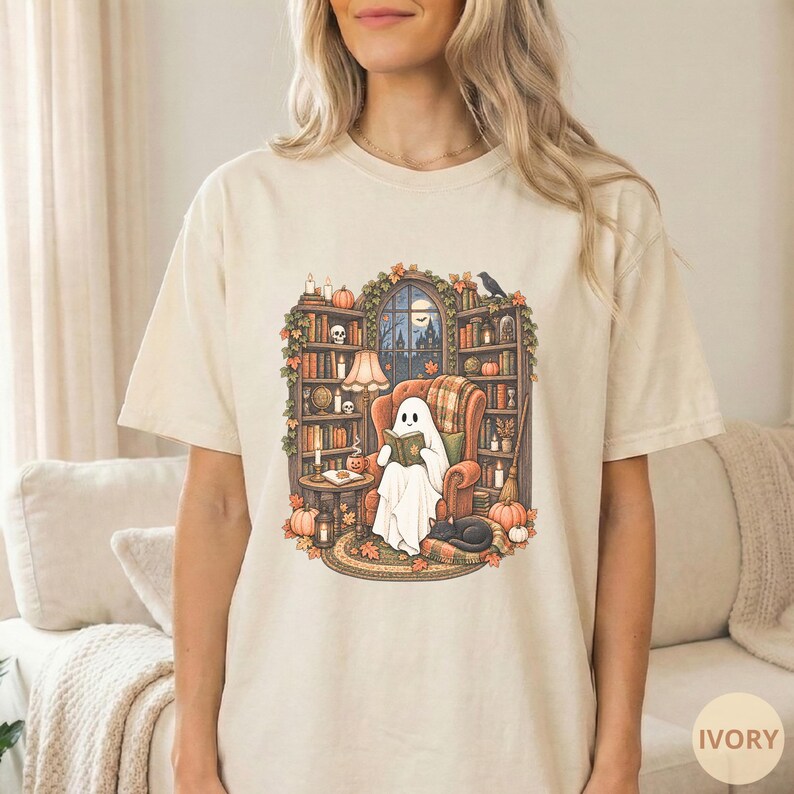 May include: An ivory short-sleeve t-shirt featuring a graphic illustration of a ghost sitting in an armchair reading a book. The scene includes bookshelves, pumpkins, a black cat, and a window with a night sky view, all rendered in warm autumn tones.