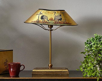 May include: A gold-toned table lamp with a rectangular base and a slender stem. The lampshade is gold with a black trim and features a painted scene of two cranes. A red mug sits to the left of the lamp.