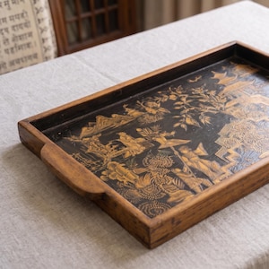 Puede incluir: Una bandeja de servicio rectangular de madera con acabado marrón oscuro y asas laterales integradas. La superficie interior presenta una base negra con intrincadas ilustraciones doradas que representan figuras, árboles, montañas y estructuras arquitectónicas.