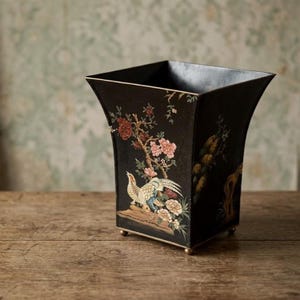 May include: Black square decorative container with a flared top and small gold feet. The container features a hand-painted design of a bird, flowers, and branches in shades of red, pink, green, and gold.