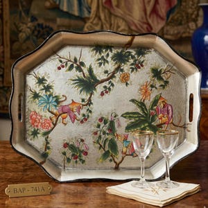 May include: An octagonal silver-toned serving tray featuring a hand-painted chinoiserie motif of monkeys in colorful clothing amidst flowering branches and berries. Two clear glass stemmed goblets sit on a folded white napkin in front of the tray.