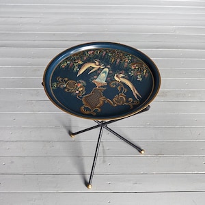 May include: An oval, dark blue serving tray with a gold rim, supported by a black metal stand. The tray features a painted design of birds, flowers, and ornate gold accents. The stand has three legs with gold-colored tips.
