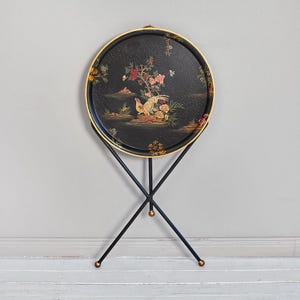 Może przedstawiać: Okrągły czarny stolik z złotą obwódką i czarnym metalowym stojakiem. Taca ma kolorowy wzór kwiatowy i ptasi. Stojak ma podstawę trójnożną ze złotymi akcentami.