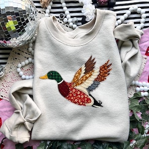 May include: Cream-colored sweatshirt featuring a colorful duck applique. The duck has a green plaid head, red floral body, and wings with brown, tan, and blue patterned fabrics. The sweatshirt is a casual, comfortable garment.