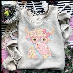 May include: A beige sweatshirt featuring a whimsical cow design. The cow has floral patches, heart-shaped glasses, and a pink bow. The cow is blowing a bubblegum bubble. The sweatshirt is a crew neck style.