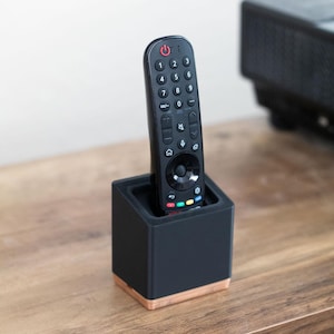 May include: A black remote control with a numeric keypad, navigation wheel, and power button, standing upright in a square black holder with a light wood base. The remote features various function icons and a red power symbol at the top.