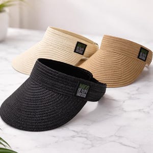 May include: Three straw visors in beige, tan, and black, each with a curved brim and a band featuring the text "NAKYO BEAUTY". The visors are arranged on a marble surface, offering sun protection.