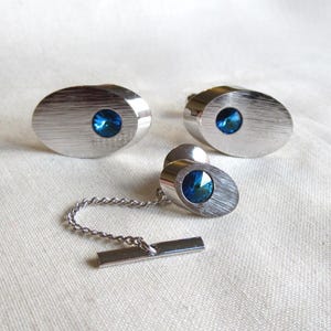 Könnte beinhalten: Ein Set aus silberfarbenen Manschettenknöpfen und einer Krawattennadel mit gebürsteter Metalloberfläche. Jedes Stück hat ein ovales Design mit einem leuchtend blauen Edelstein in der Mitte. Die Krawattennadel ist mit einer Kette und einem Steg verbunden.