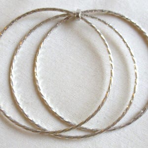Può includere: Un set di tre braccialetti rigidi color argento intrecciati tra loro. Ogni sottile fascia circolare presenta un motivo superficiale ritorto o testurizzato continuo che riflette la luce, collegati in un unico punto da un piccolo anello metallico liscio.