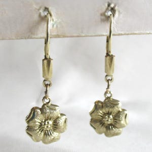 May include: A pair of gold-coloured drop earrings. Each earring features a floral design at the bottom, suspended from a lever-back closure. The earrings are displayed against a white background, highlighting their intricate details and elegant style.