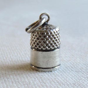 Könnte beinhalten: Ein kleiner, silberfarbener Fingerhut-Anhänger mit strukturierter Oberseite und glatter, zylindrischer Basis. Der Anhänger hat eine kleine Schlaufe zum Befestigen an einer Halskette oder einem Armband. Ein Nähzubehör.