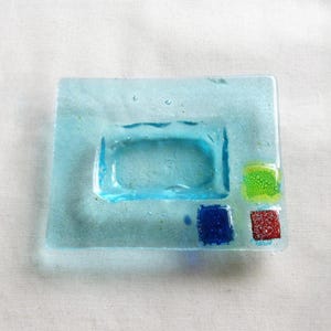 May include: A square, light blue glass dish with a rectangular indentation in the center. The dish has three colorful square accents: green, blue, and red. The glass has a textured appearance.
