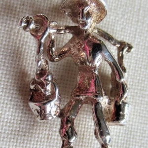 May include: Silver charm of a person carrying a bucket on their shoulder. The person is wearing a hat and has a detailed face. The charm is a 3D representation of a person.