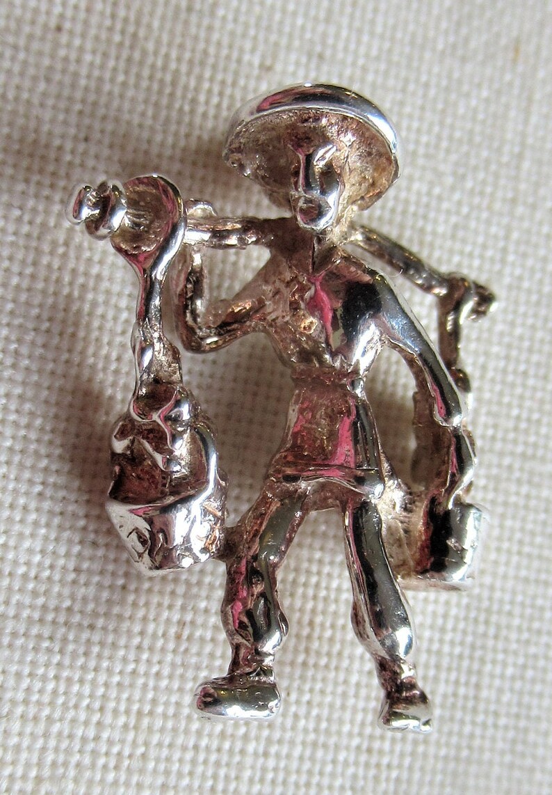 May include: Silver charm of a person carrying a bucket on their shoulder. The person is wearing a hat and has a detailed face. The charm is a 3D representation of a person.
