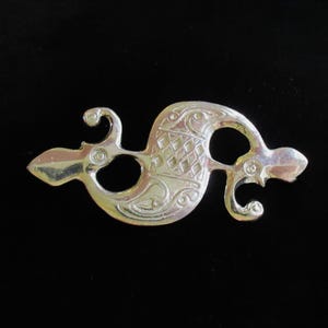 May include: A silver-colored, openwork brooch with an intricate design. The brooch features a central, diamond-patterned element, flanked by swirling, stylized shapes. The overall design suggests a historical or Celtic influence, set against a black background.