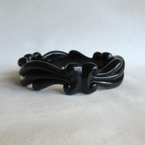 May include: A black, ornate bangle bracelet. The bracelet features a repeating, stylized floral design. The glossy, dark bracelet is made of a solid material and is set against a neutral background. This vintage-style bracelet is a fashion accessory.
