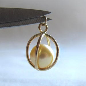 May include: A gold-toned metal pendant featuring a spherical cream-colored pearl encased within two intersecting circular gold-toned metal rings, suspended from a small circular jump ring at the top of the structure.
