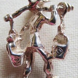 May include: Silver charm of a person carrying two bags on their back and shoulders. The charm has a loop at the top for attaching to a bracelet or necklace.