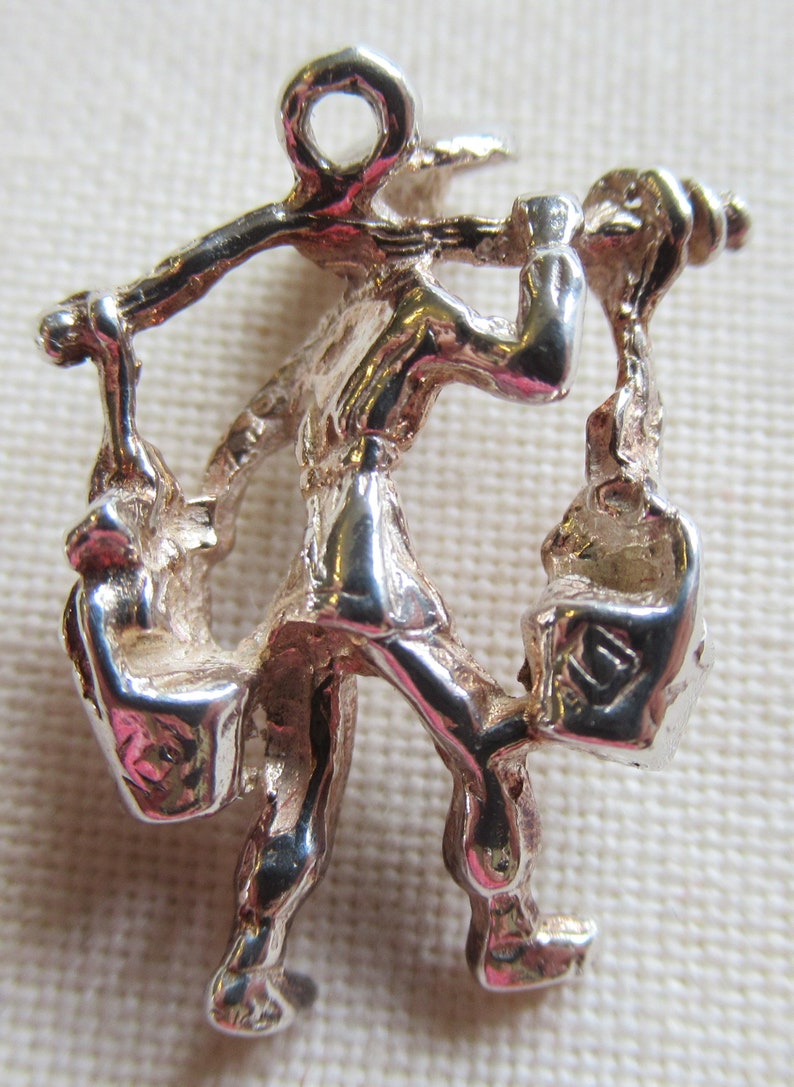 May include: Silver charm of a person carrying two bags on their back and shoulders. The charm has a loop at the top for attaching to a bracelet or necklace.