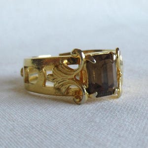 Op de afbeelding: Een goudkleurige ring met een rechthoekige, rookbruine edelsteen. De ring heeft een sierlijk ontwerp met opengewerkte filigraan- en kruldetails, wat een elegante, vintage uitstraling creëert.
