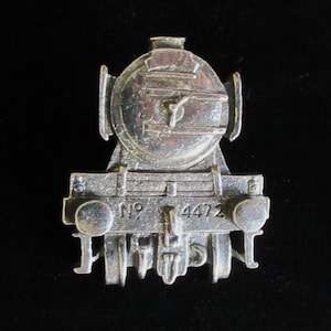 May include: A silver-colored train engine pin with the number 4472 displayed on the front. The pin is a detailed miniature of a steam locomotive, with visible wheels and a circular front. The background is black.