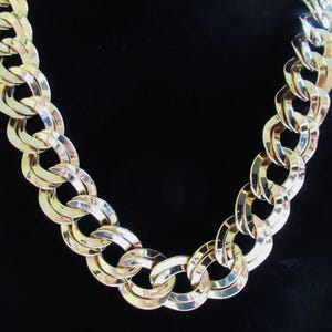 May include: A gold-coloured chain necklace featuring large, interlocking oval links. The necklace is set against a black background, emphasising its shiny, reflective surface. The chain's design is bold and eye-catching, suitable as a statement piece.