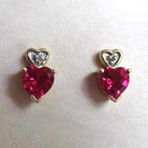 Puede incluir: Un par de pendientes de corazón de oro con una piedra roja rubí en el centro de cada corazón y una piedra más pequeña en forma de diamante en la parte superior de cada corazón.