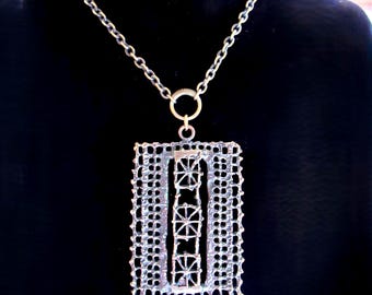 Puede incluir: Collar con colgante plateado con un diseño rectangular calado. El colgante presenta una serie de formas geométricas dentro de un marco rectangular, suspendido de una cadena. El collar se muestra sobre un fondo negro.