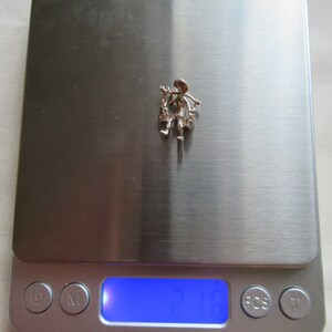 May include: A silver charm of a person carrying a basket of flowers. The charm is on a digital scale that reads 2.1 grams.