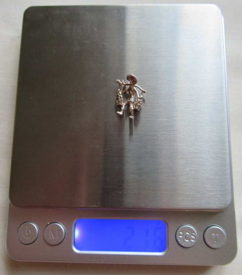 May include: A silver charm of a person carrying a basket of flowers. The charm is on a digital scale that reads 2.1 grams.