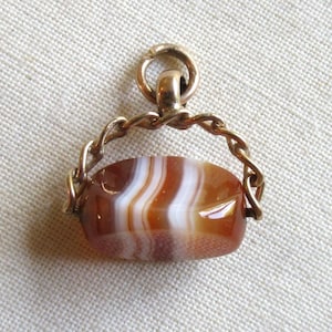 May include: A gold-toned metal swivel fob pendant featuring an oval-shaped banded agate stone with horizontal layers of translucent brown and opaque white. The stone is held within a twisted metal frame attached to a circular jump ring at the top.