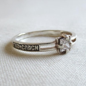 May include: A silver-toned ring featuring a clear, square-cut stone set in a four-prong mount. The band is split into two parallel lines on one side, with a rectangular black-filled engraving containing Cyrillic text integrated into the metal band.