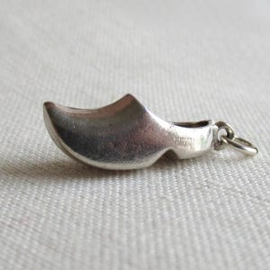 Könnte beinhalten: Ein kleiner, silberfarbener Metallanhänger in Form eines traditionellen holländischen Holzschuhs. Der Anhänger hat eine glatte, geschwungene Oberfläche und verfügt über einen kleinen runden Ring an der Ferse zum Befestigen an einer Kette.